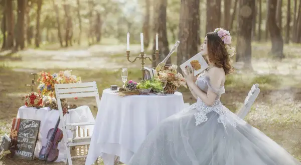 Panna młoda w sukni ślubnej czyta książkę przy stole z owocami i kwiatami. Obok skrzypce i tablica z napisem "We are getting married".