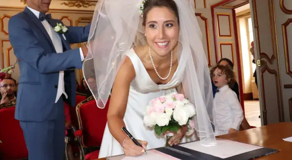 La mariée Anne-Claire Legendre signe son acte de mariage, bouquet à la main. Un enfant aux boucles blondes observe la scène.