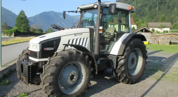 Biały traktor Hurlimann XT-130 stoi na żwirowej drodze, w tle góry i domy.