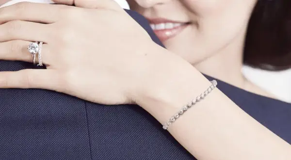 Anillo de compromiso en la mano izquierda, símbolo de amor eterno. Una mujer abraza a su pareja, luciendo un brazalete de diamantes.