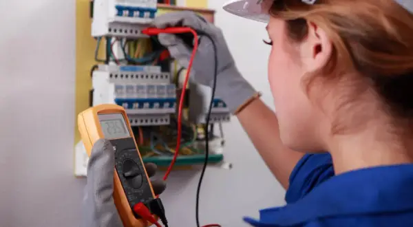Elektryk w białym kasku mierzy napięcie multimetrem przy skrzynce z bezpiecznikami.