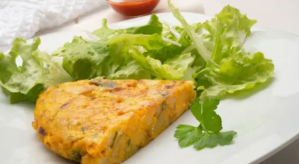 Una porción de tortilla de patatas, dorada y jugosa, acompañada de una fresca ensalada verde y un toque de perejil.