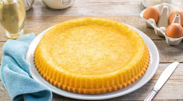Una torta furba dorata, appena sfornata, su un piatto bianco. Ingredienti come uova e olio sono visibili sullo sfondo.