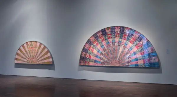 Two fan-shaped artworks display intricate pattern and decoration, creating a sense of movement. The larger piece bursts with colorful fabric squares, while the smaller one features a softer, sunburst design.