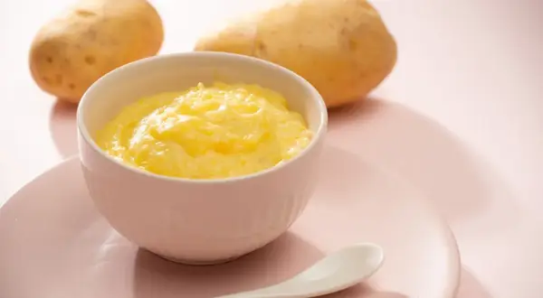 Ein Schälchen mit cremigem Kartoffelbrei, bereit für ein Baby. Zwei Kartoffeln liegen im Hintergrund.