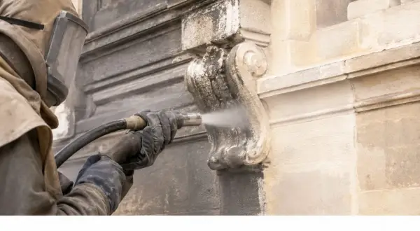 Restauración de fachadas con limpieza a chorro de arena. Un operario con equipo de protección limpia detalles ornamentales de piedra.