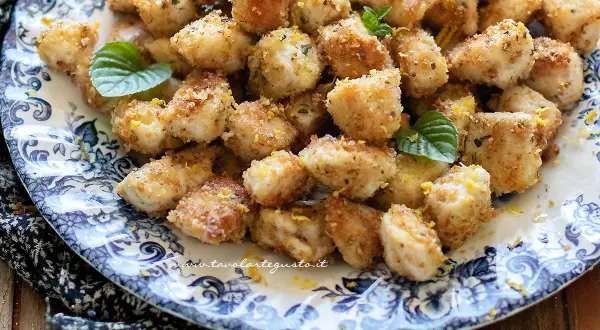 Bocconcini di pollo dorati e croccanti, guarniti con scorza di limone e foglie di menta fresca su un piatto decorato.