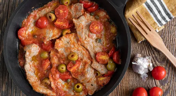 Thinly sliced meat in a tomato sauce with olives and cherry tomatoes in a pan.