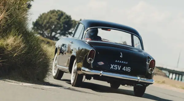 Svart Simca Aronde glider längs en kustväg, en klassisk skönhet bland gräs och träd.