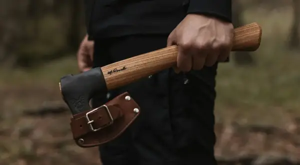 Mano sujetando hachas tácticas con funda de cuero. El mango de madera tiene una inscripción.