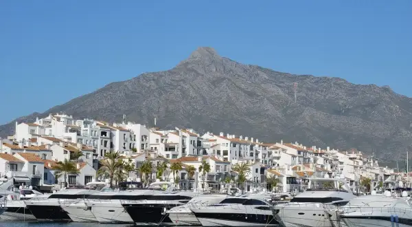 Luksusowe jachty cumują w malowniczym porcie Puerto Banús, z białymi budynkami i górą w tle.