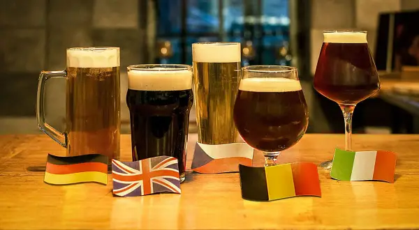 Cinque bicchieri di birre scure famose, ognuno con una bandiera nazionale: Germania, Regno Unito, Repubblica Ceca, Belgio e Italia.