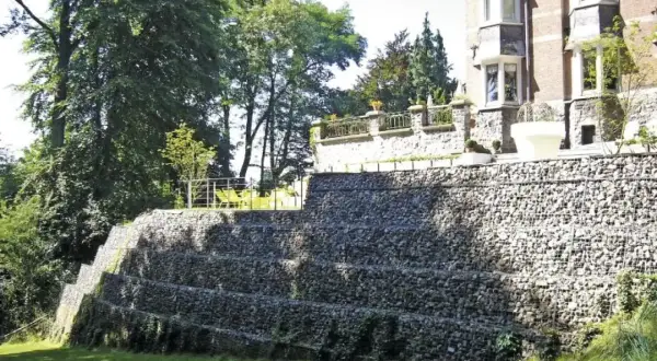 Imponująca skarpa na granicy działki, zbudowana z kamieni w gabionach, tworzy tarasy porośnięte zielenią. Na górze widać fragment rezydencji.