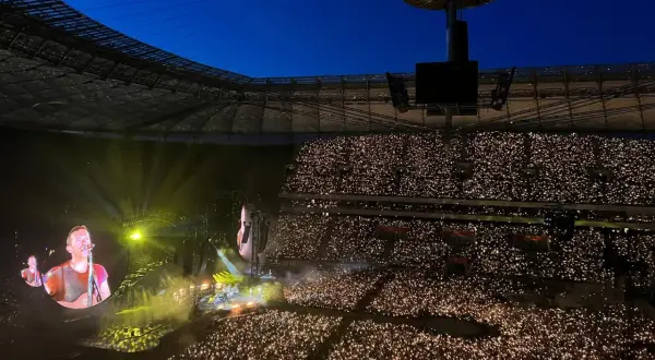 Koncert na stadionie, tłum z zapalonymi telefonami, na ekranie wokalista Coldplay.