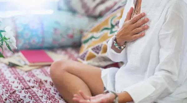 Frau in weißer Kleidung praktiziert buddhistische Herzmeditation, Hände auf der Brust und im Schoß, umgeben von Kissen und Büchern.