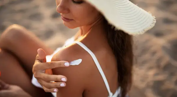 Kobieta w kapeluszu smaruje ramię kremem z filtrem SPF 50, chroniąc skórę przed słońcem na plaży.