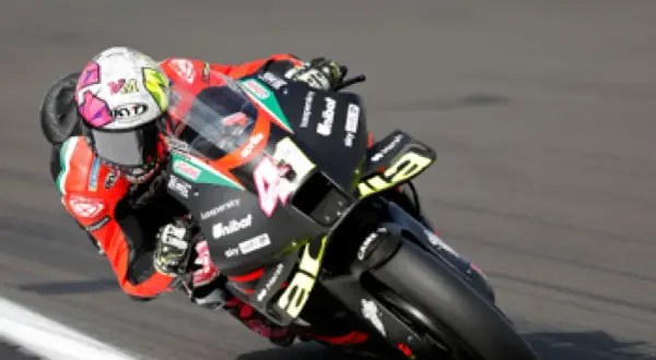 Piloto de MotoGP inclinado en una curva, con su casco blanco y rosa y la moto negra con detalles amarillos.