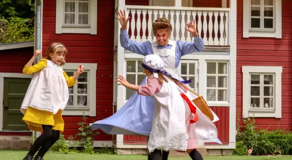 Barn leker vid Junibacken Madicken hus. En vuxen i blå klänning och två barn i tidsenliga kläder.