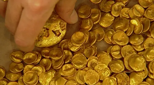 Ein Finger berührt einen Teil des Goldschatzes von Manching, der aus vielen kleinen, unregelmäßigen Goldmünzen besteht.