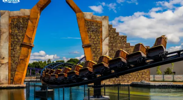 Ekipa rollercoastera w Energylandii gotowa na przygodę! Drewniane wagoniki pędzą nad wodą, a za nimi widać fioletowe tory.