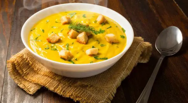 Un fumante passato di verdure giallo brillante, guarnito con crostini dorati e erbe fresche, servito in una ciotola bianca su una tovaglietta di juta.