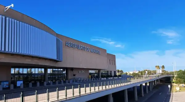 Nowoczesny budynek lotniska Aeropuerto de Menorca z napisem "AEROPUERTO DE MENORCA" i podjazdem pod niebo.