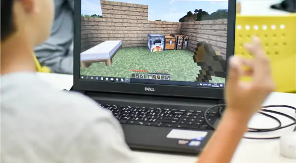 Chłopiec gra w Minecrafta na laptopie, ucząc się jak dodać mody na Aternos. Widok na ekranie gry z łóżkiem, piecem i skrzynią.