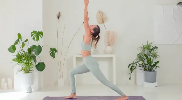 Donna in posa yoga, che mostra i benefici di una pratica regolare. L'ambiente è luminoso e pieno di piante, evocando calma e benessere.