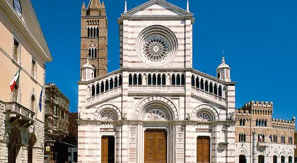 Il Duomo di Grosseto, con la sua facciata a strisce bianche e rosa, domina la piazza. Accanto, un campanile medievale si staglia contro il cielo azzurro.