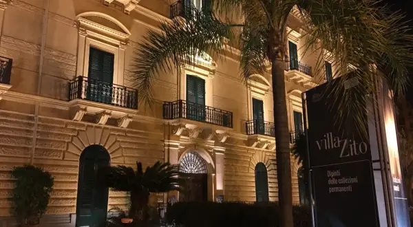 Villa Zito a Palermo, un'elegante dimora illuminata di notte, con palme che incorniciano la facciata e un cartello che annuncia "Dipinti delle collezioni permanenti".