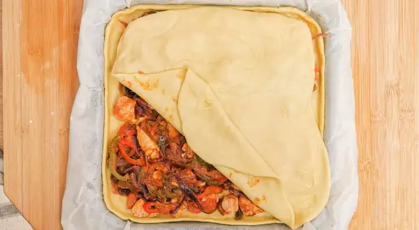 Empanada de pollo lista para hornear, con relleno de pollo y verduras cubierto por masa dorada.