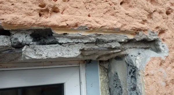 Uszkodzony montaż nadproża w starym domu, widoczne zbrojenie i luźne elementy betonowe nad oknem.