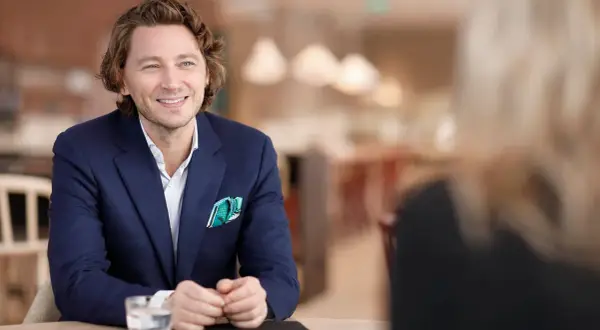 Rasmus Nerman, klädd i en mörkblå kavaj, ler och tittar åt sidan under ett samtal vid ett bord.