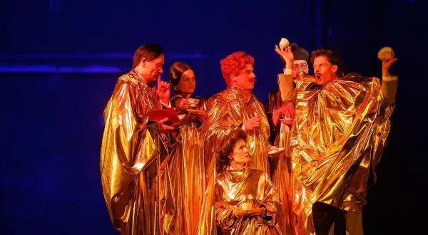 Aktorzy w złotych szatach, trzymający filiżanki i książki, tworzą scenę z najlepszych spektakli teatralnych.