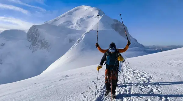 Un alpiniste victorieux lève ses bâtons sur fond de Mont Blanc. L'ascension est réussie, la neige brille sous le ciel bleu.