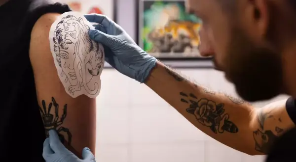 Artysta w rękawiczkach dopasowuje wzór tatuażu na ramieniu klienta. Widać już kilka gotowych tatuaży, co sugeruje, że to miejsce na tatuaż jest popularne.