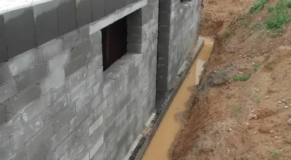 Budowa fundamentów z bloczków betonowych, obok wykop z wodą, który wymaga drenażu.