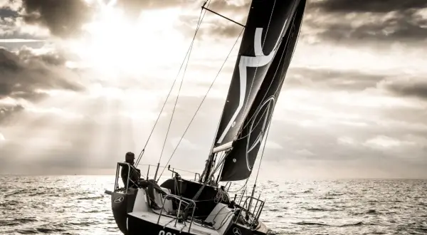 Vela nera con logo, un uomo a bordo di una barca a vela che naviga in mare con un cielo drammatico. L'imbarcazione mostra un angolo di deriva accentuato.