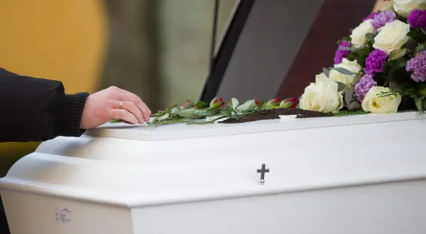 Dłoń kładzie róże na białej trumnie ozdobionej kwiatami i krzyżem.