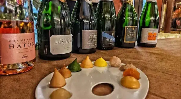 Bottiglie di champagne, tra cui Veuve Clicquot e Bruno Paillard, disposte su un tavolo con una tavolozza da pittore piena di dolcetti colorati, un'ode al gusto e all'arte del cote de bar.