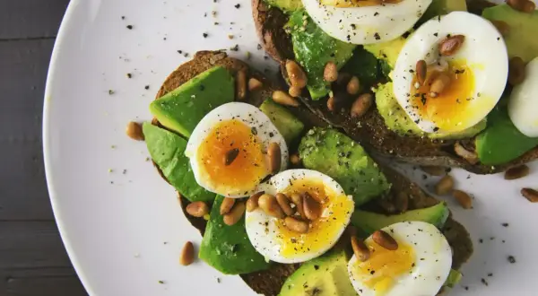 Avokado recept: två rostade brödskivor med avokado, kokt ägg och pinjenötter, strösslade med peppar.