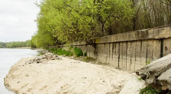 Betonowa opaska na rzece chroni brzeg przed erozją. Wiosenne drzewa tworzą zieloną ścianę nad wodą.