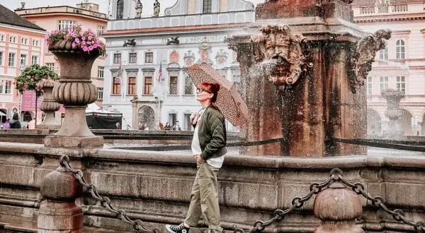 Regenschirm-Schwung am Brunnen in Budweis Stadt. Die Frau genießt die frische Brise, während die historische Kulisse im Hintergrund erstrahlt.
