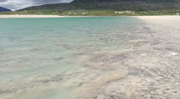 Vackert turkost vatten möter en sandstrand med mönster från tidvattnet. Hus och en grön kulle syns i bakgrunden på Yttre Hebriderna.
