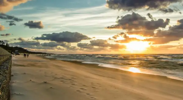 Zachód słońca nad Bałtykiem, plaża i falochron. W oddali widać ludzi spacerujących i kitesurferów. To idealne miejsce na podziwianie atrakcji Jastrzębiej Góry i okolic.