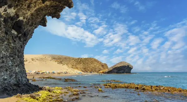 Vista de la playa de San José con rocas cubiertas de algas en primer plano y gente disfrutando del sol bajo sombrillas.
