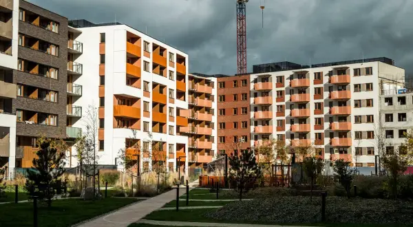 Nowe bloki mieszkalne z balkonami i żurawiem budowlanym w tle. Widać zielony dziedziniec, ścieżki i drzewa. Rynek wtórny w fazie rozwoju.