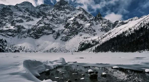 Morskie Oko zimową porą. Majestatyczne, ośnieżone szczyty Tatr górują nad zamarzniętym jeziorem. W oddali widać ludzi spacerujących po lodzie.