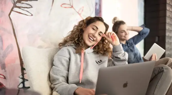 Zwei junge Frauen lernen entspannt. Eine trägt einen Hoodie mit dem Logo der IU Internationale Hochschule, die andere hält ein Tablet.