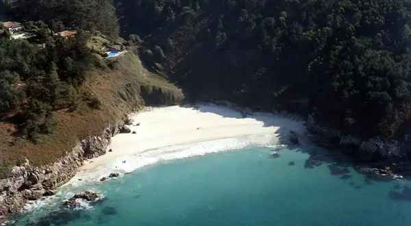 Vista aérea de la playa de San Pedro, Galicia, con aguas turquesas, arena blanca y vegetación exuberante en los acantilados.